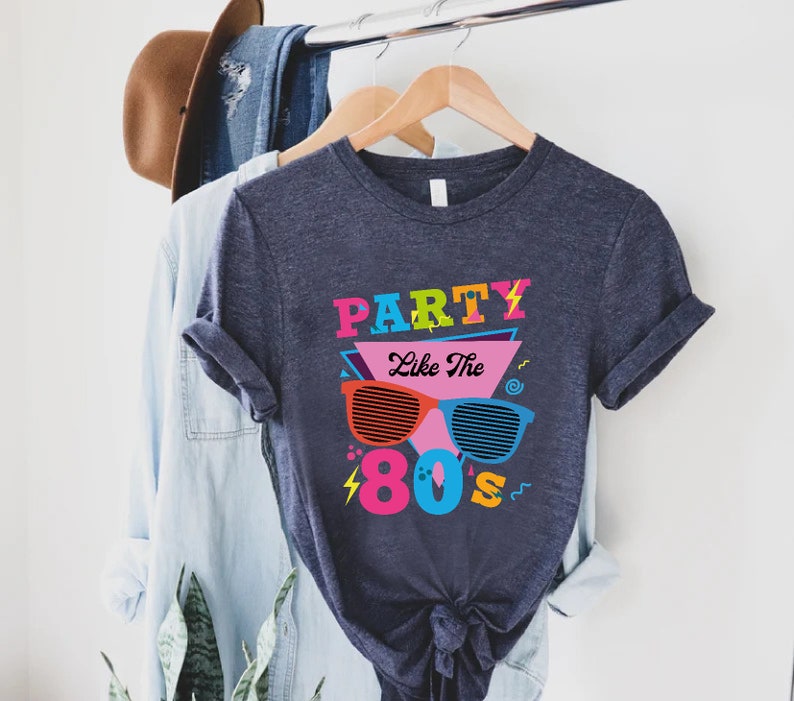 K&ouml;nnte beinhalten: Ein dunkelblaues T-Shirt mit einem grafischen Design, das den Text "Party Like The 80's" in bunten Buchstaben und eine Retro-Sonnenbrille zeigt.
