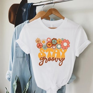 May include: White t-shirt with a graphic print of flowers and peace signs. The text "Stay groovy" is printed in yellow and brown letters.