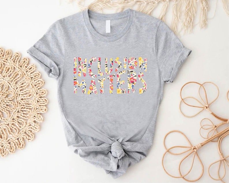 Puede incluir: Una camiseta gris con el texto "Inclusion Matters" impreso en letras florales.