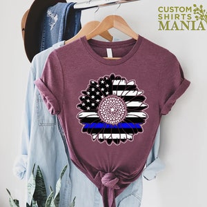 May include: A maroon t-shirt featuring a sunflower design. The sunflower is made up of the American flag with a blue line running through the centre.