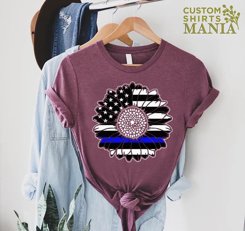 May include: A maroon t-shirt featuring a sunflower design. The sunflower is made up of the American flag with a blue line running through the centre.