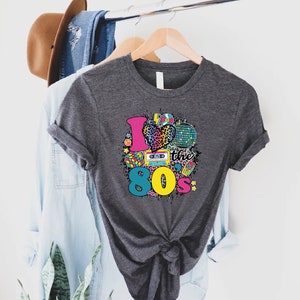 May include: A dark gray t-shirt with a graphic print of a heart with a leopard print design and the text "I love the 80s" surrounded by 80s themed icons.