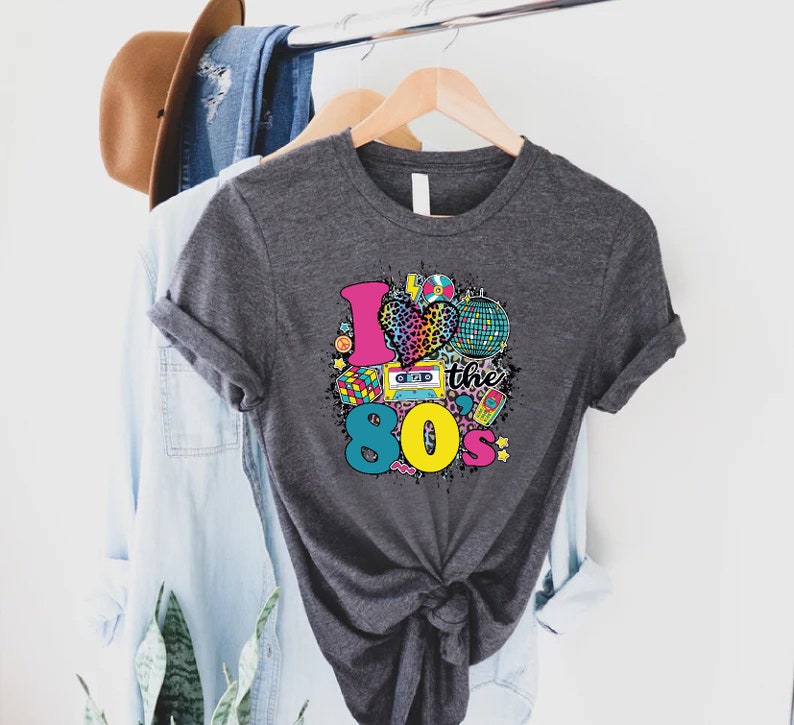 May include: A dark gray t-shirt with a graphic print of a heart with a leopard print design and the text "I love the 80s" surrounded by 80s themed icons.