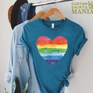 Peut inclure: T-shirt bleu sarcelle avec un motif de c&oelig;ur arc-en-ciel &agrave; l'aquarelle. Le t-shirt est suspendu &agrave; un cintre en bois. Le texte "CUSTOM SHIRTS MANIA" est visible en haut &agrave; droite. D'autres v&ecirc;tements et un chapeau sont &eacute;galement visibles.