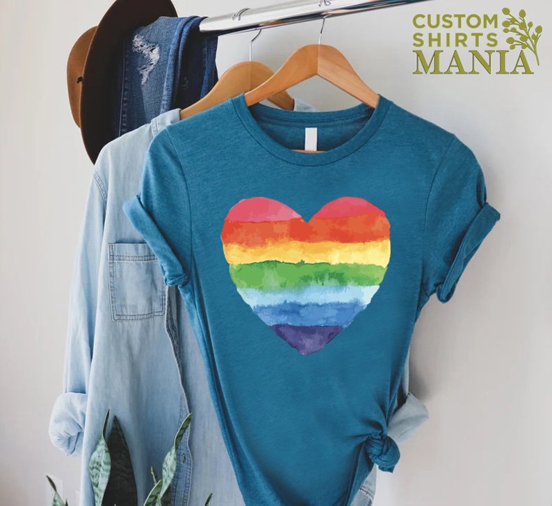 Peut inclure: T-shirt bleu sarcelle avec un motif de c&oelig;ur arc-en-ciel &agrave; l'aquarelle. Le t-shirt est suspendu &agrave; un cintre en bois. Le texte "CUSTOM SHIRTS MANIA" est visible en haut &agrave; droite. D'autres v&ecirc;tements et un chapeau sont &eacute;galement visibles.