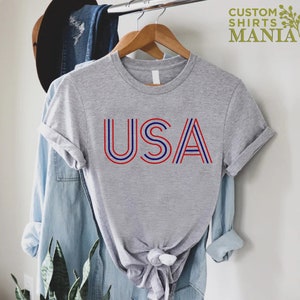 Może przedstawiać: Szara koszulka z nadrukiem "USA" w czerwono-niebieskie paski. Koszulka jest na wieszaku i ma zawiązany d&oacute;ł.