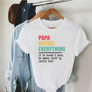 May include: White t-shirt with the text "PAPA KNOWS EVERYTHING" in red, yellow, and teal. Below the text is the phrase "If he doesn't know, he makes stuff up really fast". The shirt is hanging on a wooden hanger.