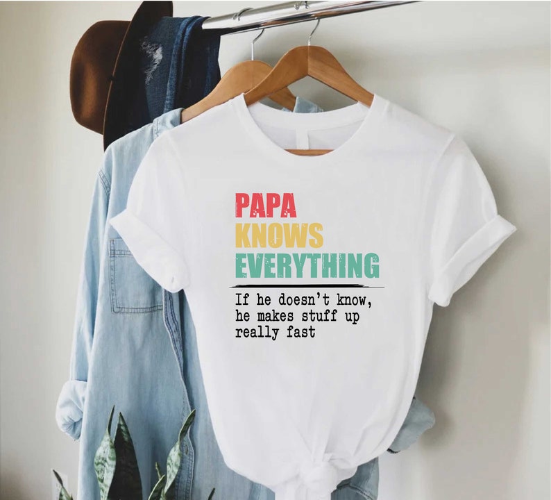 May include: White t-shirt with the text "PAPA KNOWS EVERYTHING" in red, yellow, and teal. Below the text is the phrase "If he doesn't know, he makes stuff up really fast". The shirt is hanging on a wooden hanger.