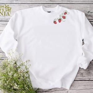 Puede incluir: Una sudadera blanca con cuello redondo, con un dise&ntilde;o bordado de fresas rojas con vides verdes. El texto "CUSTOM SHIRTS MANIA" es visible en la esquina superior izquierda. La sudadera se muestra sobre una superficie de madera.