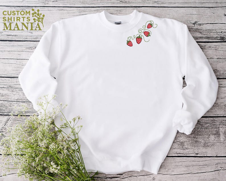 Puede incluir: Una sudadera blanca con cuello redondo, con un dise&ntilde;o bordado de fresas rojas con vides verdes. El texto "CUSTOM SHIRTS MANIA" es visible en la esquina superior izquierda. La sudadera se muestra sobre una superficie de madera.