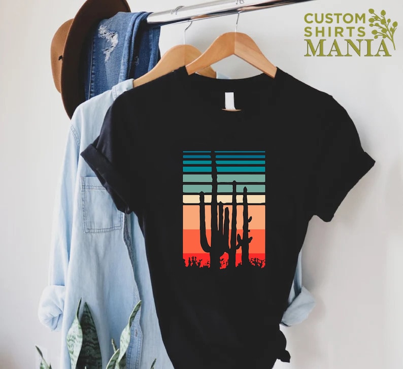 May include: Black t-shirt with a retro sunset graphic featuring three silhouette cacti. The sunset is in shades of orange, red, yellow, blue, and green.
