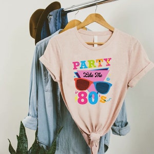K&ouml;nnte beinhalten: Ein pinkfarbenes T-Shirt mit einem grafischen Design, auf dem "Party Like The 80's" steht, mit einer Sonnenbrille im Design.