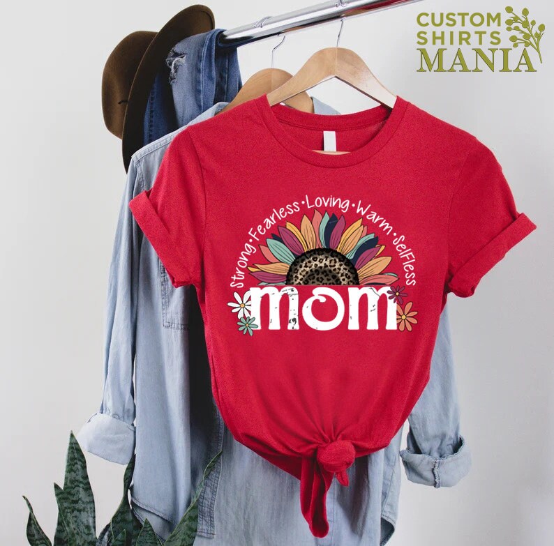 May include: Red t-shirt featuring a white graphic of a sunflower with a leopard print centre and the word "mom" in white with flowers around it. The text around the sunflower reads "Strong, Fearless, Loving, Warm, Selfless".