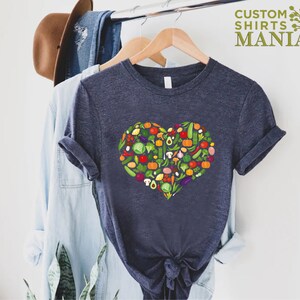 May include: Navy blue t-shirt featuring a heart-shaped design composed of various colorful vegetables. The shirt is hanging on a wooden hanger with other clothing items. The text "CUSTOM SHIRTS MANIA" is printed in green.