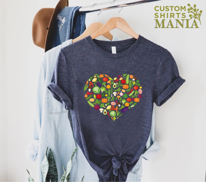 May include: Navy blue t-shirt featuring a heart-shaped design composed of various colorful vegetables. The shirt is hanging on a wooden hanger with other clothing items. The text "CUSTOM SHIRTS MANIA" is printed in green.