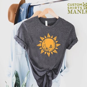 Puede incluir: Una camiseta gris oscuro con un gr&aacute;fico de sol amarillo que presenta una mariposa y detalles florales. El texto "CUSTOM SHIRTS MANIA" es visible en la esquina superior derecha de la imagen.