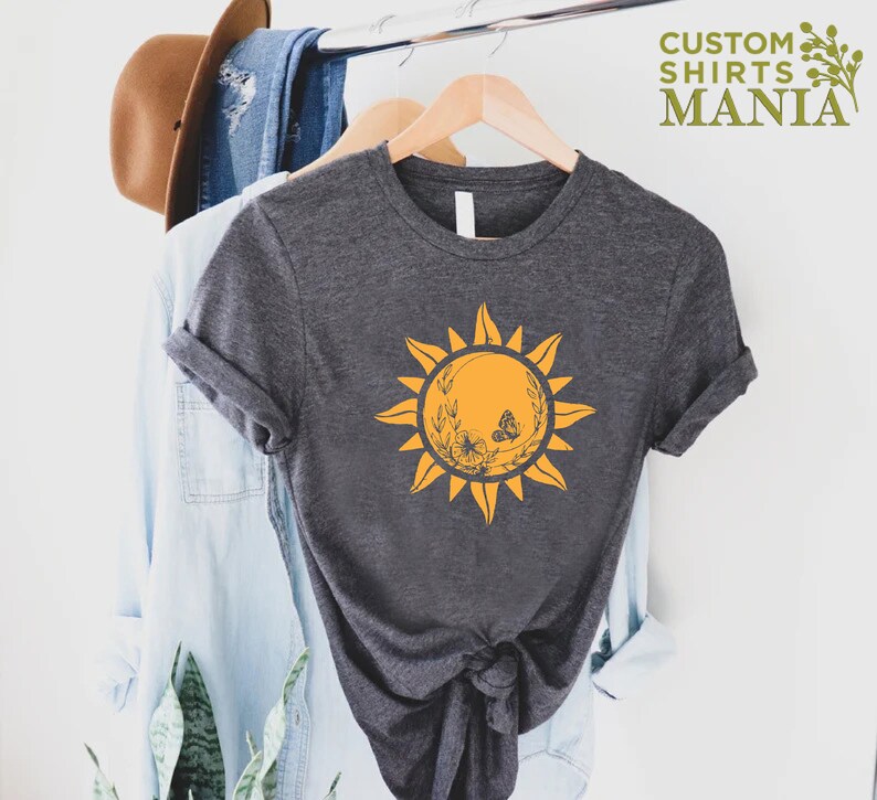 Puede incluir: Una camiseta gris oscuro con un gr&aacute;fico de sol amarillo que presenta una mariposa y detalles florales. El texto "CUSTOM SHIRTS MANIA" es visible en la esquina superior derecha de la imagen.
