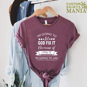 May include: A maroon t-shirt with the text "I'm going to let... God fix it because if I fix it I'm going to jail".