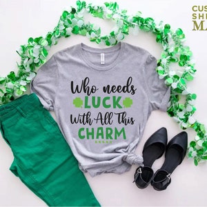 May include: A gray t-shirt with the text "Who needs luck with all this charm" and a shamrock graphic. The shirt is laid out on a white surface with green garland and black shoes.