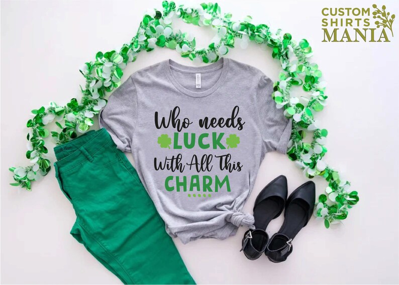 May include: A gray t-shirt with the text "Who needs luck with all this charm" and a shamrock graphic. The shirt is laid out on a white surface with green garland and black shoes.
