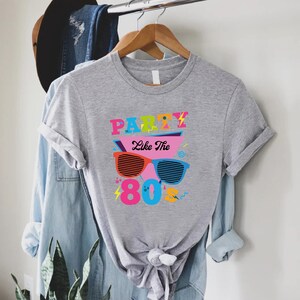 K&ouml;nnte beinhalten: Graues T-Shirt mit einem grafischen Design mit dem Text "Party Like The 80's" in bunten Buchstaben und einer Sonnenbrille im Retro-Design.