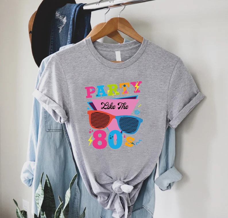K&ouml;nnte beinhalten: Graues T-Shirt mit einem grafischen Design mit dem Text "Party Like The 80's" in bunten Buchstaben und einer Sonnenbrille im Retro-Design.