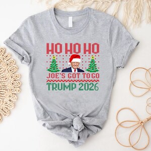 Op de afbeelding: Een grijs gem&ecirc;leerd t-shirt met een grafische print met de tekst HO HO HO, JOE'S GOT TO GO en TRUMP 2026. Het ontwerp bevat een centrale afbeelding van Donald Trump met een kerstmuts, geflankeerd door twee groene kerstbomen, alles in een rood, groen en wit breipatroon.