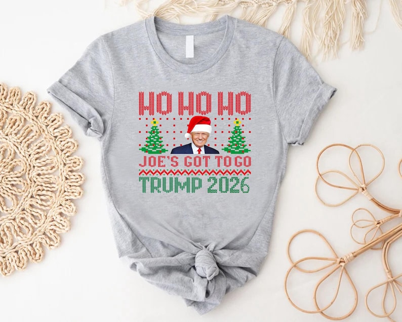 Op de afbeelding: Een grijs gem&ecirc;leerd t-shirt met een grafische print met de tekst HO HO HO, JOE'S GOT TO GO en TRUMP 2026. Het ontwerp bevat een centrale afbeelding van Donald Trump met een kerstmuts, geflankeerd door twee groene kerstbomen, alles in een rood, groen en wit breipatroon.