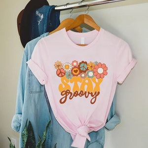 May include: A pink t-shirt with a graphic of colorful flowers and peace signs. The text "Stay groovy" is printed in a retro font.