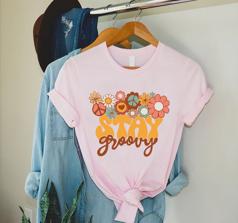 May include: A pink t-shirt with a graphic of colorful flowers and peace signs. The text "Stay groovy" is printed in a retro font.