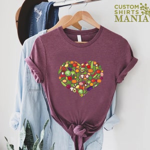 May include: Burgundy t-shirt with a heart-shaped design filled with colorful vegetables. The vegetables include tomatoes, peppers, mushrooms, and more. The text "CUSTOM SHIRTS MANIA" is in the upper right corner.