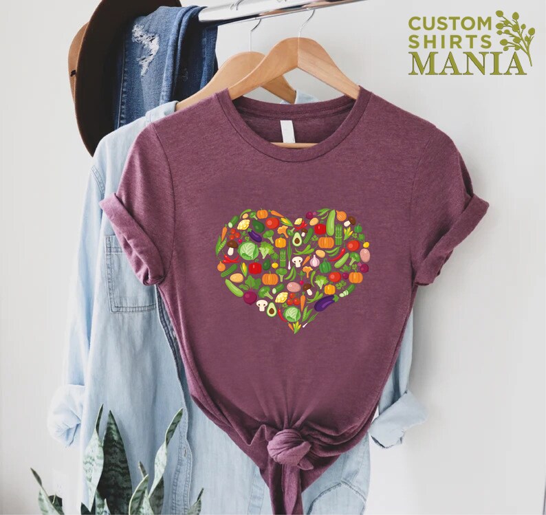 May include: Burgundy t-shirt with a heart-shaped design filled with colorful vegetables. The vegetables include tomatoes, peppers, mushrooms, and more. The text "CUSTOM SHIRTS MANIA" is in the upper right corner.