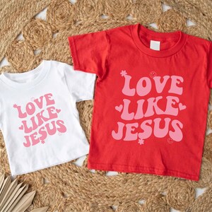 Może przedstawiać: Dwie białe koszulki z r&oacute;żowym napisem "Love Like Jesus" na czerwono-białym tle.