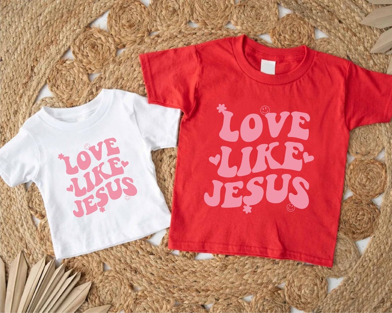 Może przedstawiać: Dwie białe koszulki z r&oacute;żowym napisem "Love Like Jesus" na czerwono-białym tle.