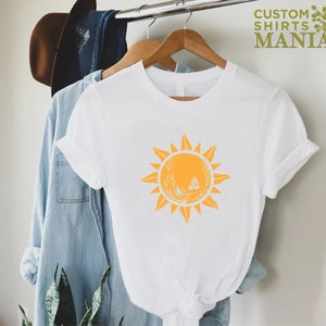 Puede incluir: Camiseta blanca con un gr&aacute;fico de sol amarillo con una mariposa y detalles florales. El texto "CUSTOM SHIRTS MANIA" es visible en la esquina superior derecha de la imagen.