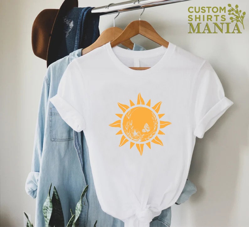 Puede incluir: Camiseta blanca con un gr&aacute;fico de sol amarillo con una mariposa y detalles florales. El texto "CUSTOM SHIRTS MANIA" es visible en la esquina superior derecha de la imagen.