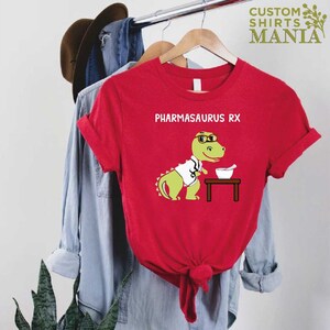 May include: Red t-shirt featuring a green dinosaur wearing a white lab coat and glasses. The dinosaur is holding a mortar and pestle. The text "PHARMASAURUS RX" is printed on the shirt.