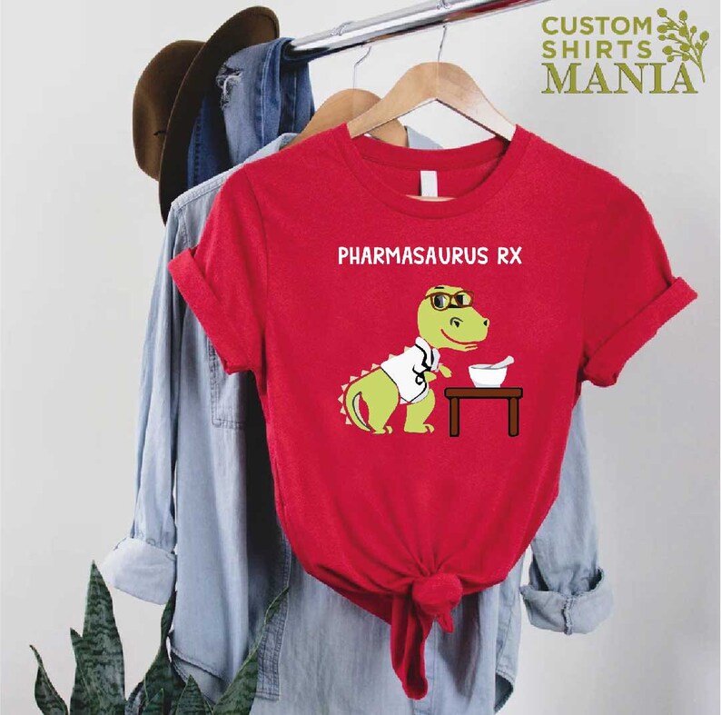 May include: Red t-shirt featuring a green dinosaur wearing a white lab coat and glasses. The dinosaur is holding a mortar and pestle. The text "PHARMASAURUS RX" is printed on the shirt.