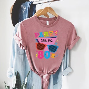 K&ouml;nnte beinhalten: Ein pinkfarbenes T-Shirt mit dem Text "Party Like The 80's" in bunten Buchstaben und einer Grafik von Retro-Sonnenbrillen.