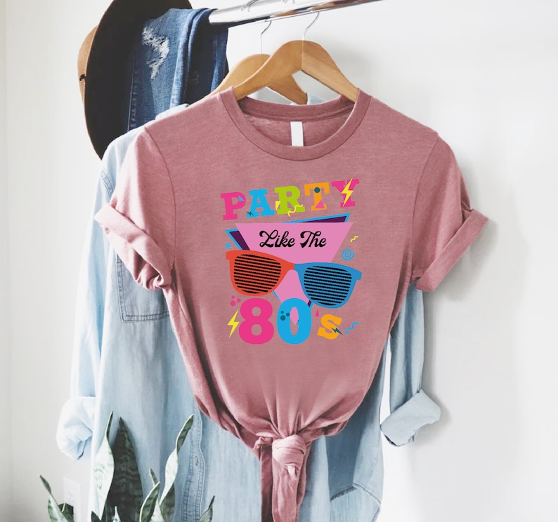 K&ouml;nnte beinhalten: Ein pinkfarbenes T-Shirt mit dem Text "Party Like The 80's" in bunten Buchstaben und einer Grafik von Retro-Sonnenbrillen.