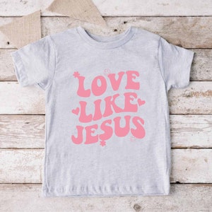 Może przedstawiać: Jasnoszary t-shirt z r&oacute;żowym nadrukiem, na kt&oacute;rym widnieje napis "LOVE LIKE JESUS" w retro czcionce. Nadruk zawiera r&oacute;wnież małe r&oacute;żowe serduszka i gwiazdki.