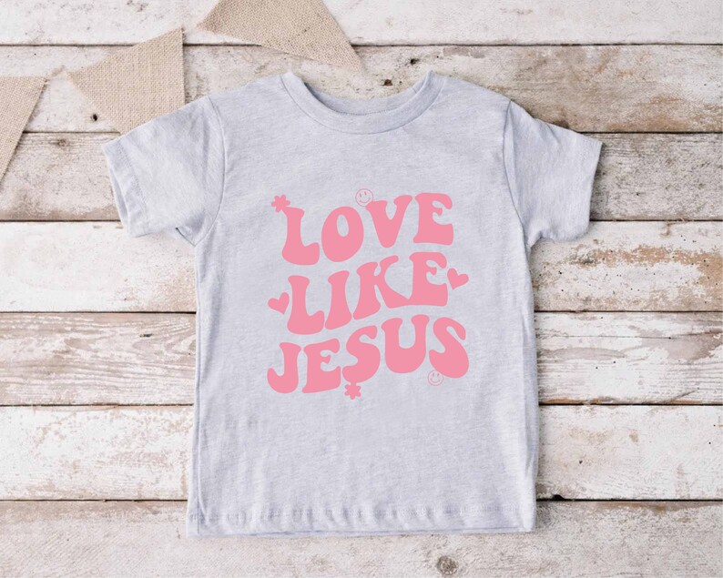 Może przedstawiać: Jasnoszary t-shirt z r&oacute;żowym nadrukiem, na kt&oacute;rym widnieje napis "LOVE LIKE JESUS" w retro czcionce. Nadruk zawiera r&oacute;wnież małe r&oacute;żowe serduszka i gwiazdki.
