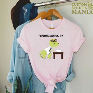 May include: Pink t-shirt featuring a cartoon dinosaur wearing glasses and a lab coat. The dinosaur is holding a bowl and a spoon. The text "PHARMASAURUS RX" is printed on the shirt.