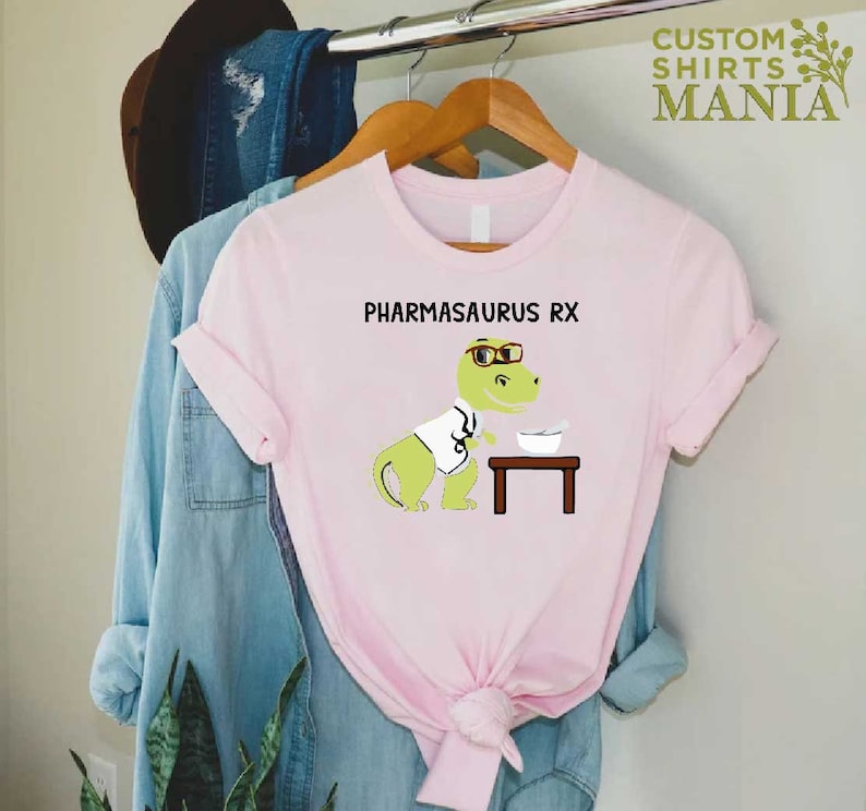 May include: Pink t-shirt featuring a cartoon dinosaur wearing glasses and a lab coat. The dinosaur is holding a bowl and a spoon. The text "PHARMASAURUS RX" is printed on the shirt.