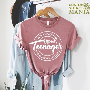 May include: A pink t-shirt with a white circle graphic that says "Warning Official Teenager May Contain Attitude".