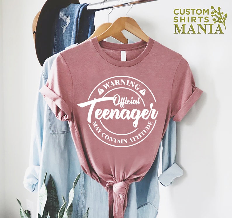 May include: A pink t-shirt with a white circle graphic that says "Warning Official Teenager May Contain Attitude".