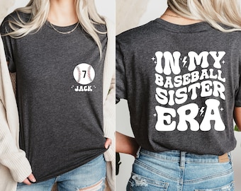 May include: A dark gray t-shirt with a white baseball graphic and the text "7 JACK" on the front. The back of the shirt has white text that reads "IN MY BASEBALL SISTER ERA" with lightning bolts.