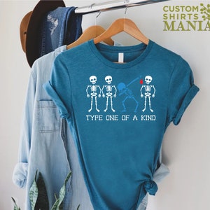 以下が含まれることがあります： ターコイズブルーのTシャツに、4つの骸骨のグラフィックがプリントされています。3つの骸骨は一列に並んで立っており、4つ目の骸骨はダブをしています。「Type One of a Kind」というテキストが骸骨の下にプリントされています。