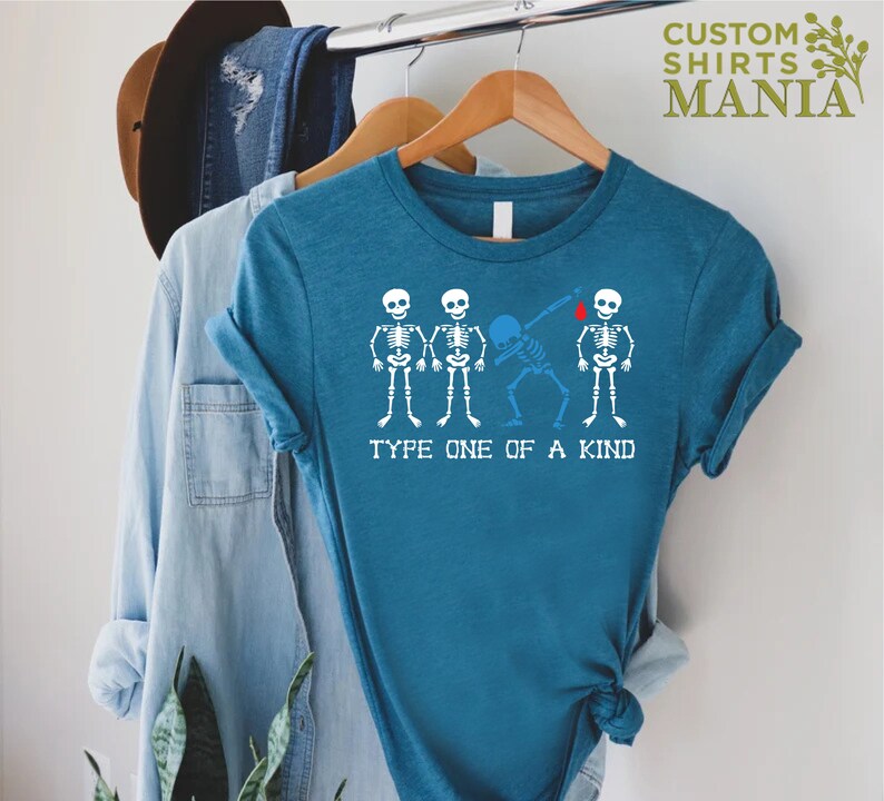 以下が含まれることがあります： ターコイズブルーのTシャツに、4つの骸骨のグラフィックがプリントされています。3つの骸骨は一列に並んで立っており、4つ目の骸骨はダブをしています。「Type One of a Kind」というテキストが骸骨の下にプリントされています。