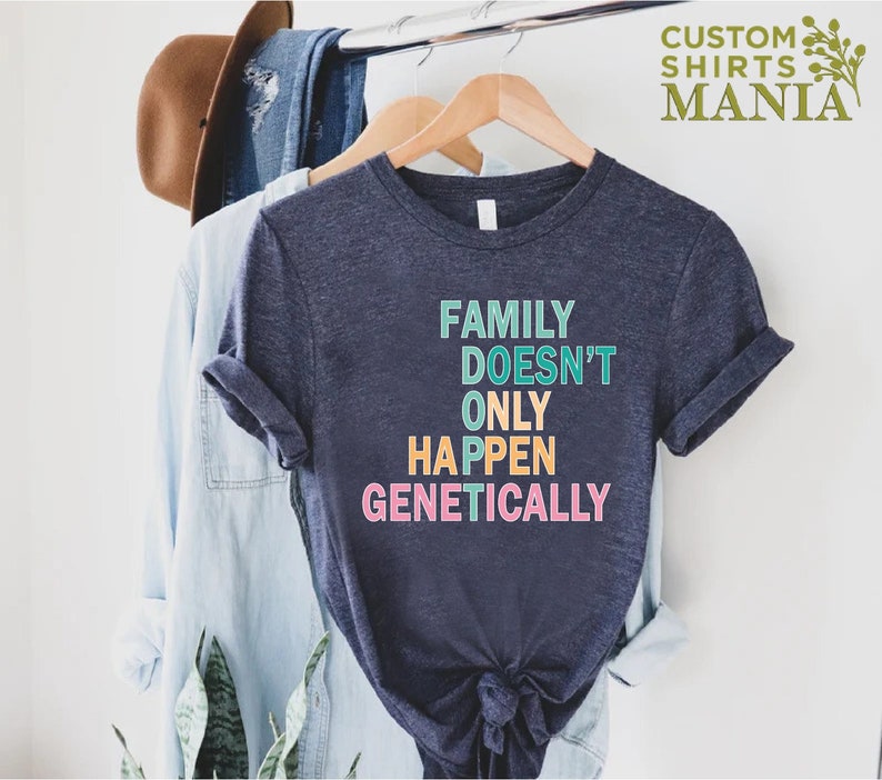 Op de afbeelding: Een marineblauw t-shirt met de tekst "FAMILY DOESN'T ONLY HAPPEN GENETICALLY" in kleurrijke letters. Het shirt hangt aan een houten hanger.